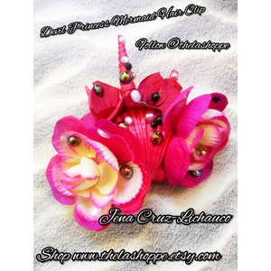 Puede incluir: Una pinza para el pelo hecha a mano con temática de sirena. La pinza presenta flores de tela rosa brillante y amarilla, adornadas con cuentas y conchas iridiscentes. El texto en la imagen dice "Devil Princess Mermaid Hair Clip" y "Follow @thelashoppe".