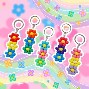 May include: Five colorful beaded keychains in a flower design. Each keychain features a different color scheme, including red, orange, yellow, green, blue, and purple. The keychains are attached to silver key rings and are set against a pink background.