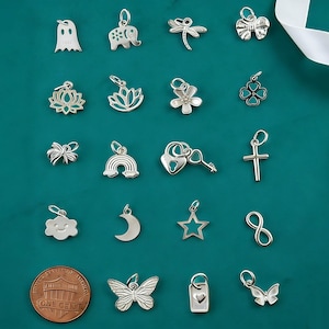 May include: A collection of twenty small silver-toned metal charms featuring various shapes including a ghost, elephant, dragonfly, bow, lotus flowers, clover, rainbow, heart lock with key, cross, cloud, moon, star, infinity symbol, butterfly, and tag.