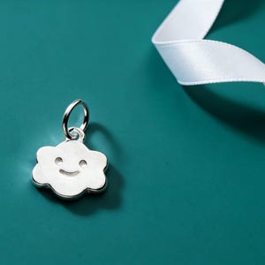 May include: A silver cloud charm with a smiling face, suspended from a small loop. The charm is set against a teal background with a white ribbon. The charm is approximately 2 cm wide.