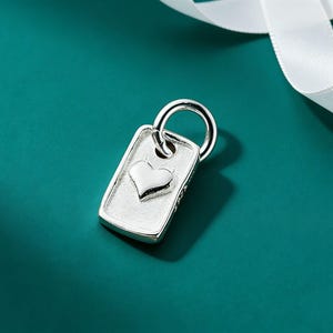 May include: A silver-colored rectangular pendant with a heart-shaped design. The pendant has a small ring at the top for attachment. A white ribbon is in the background. The pendant is likely made of metal.