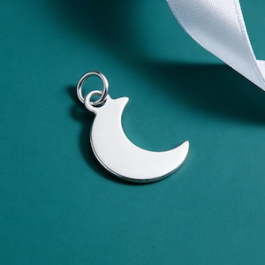 May include: A silver crescent moon charm with a small loop for attaching to a necklace or bracelet. The charm is set against a teal background with a white ribbon in the upper right corner.