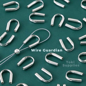Peut inclure: Une collection de protège-fils en forme de U de couleur argentée dispersés sur une surface vert foncé. Un protège-fil est enfilé avec un fil argenté fin et une petite perle ronde. Le texte Wire Guardian et Toki Supplies est visible.