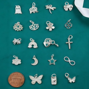 May include: A collection of twenty small silver-toned metal charms featuring various shapes including a ghost, elephant, dragonfly, bow, lotus, flower, clover, rainbow, heart lock with key, cross, cloud, moon, star, infinity symbol, butterfly, and tag.