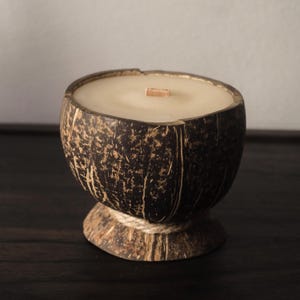 May include: A candle in a natural coconut shell. The candle has a light beige wax and a wooden wick. The coconut shell is dark brown with a textured surface and sits on a pedestal base wrapped with twine.