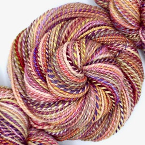 May include: Close-up of colorful yarn skeins. The yarn is a mix of pink, purple, yellow, and orange hues, creating a vibrant and textured appearance. The yarn is tightly wound, showcasing the intricate details of the fibers. Ideal for knitting and crochet projects.