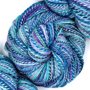 May include: Close-up of several skeins of colorful yarn. The yarn is a mix of blues, purples, and white, creating a textured, variegated effect. The yarn is tightly wound, showing the individual strands. Ideal for knitting and crochet projects.