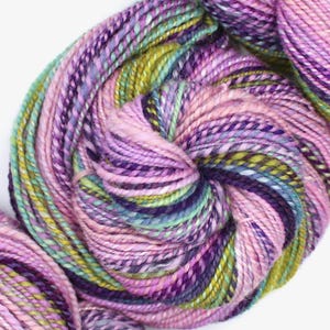 May include: A close-up of a skein of yarn in shades of pink, purple, green, and blue. The yarn is tightly wound, showcasing a blend of colors and textures, ideal for knitting or crocheting projects.