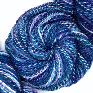 May include: Close-up of hand-dyed yarn in shades of blue, purple, and white. The yarn is tightly wound into a skein, showcasing the color variations. Ideal for knitting, crocheting, or other fiber arts projects.