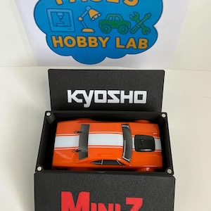May include: An orange and white Mini-Z model car inside a black box. The box has the word "Mini-Z" in red letters. The top of the box has the word "Kyosho" in white letters. The image also shows a blue and yellow sign that says "Paul's Hobby Lab".