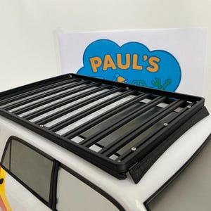 May include: A black roof rack is mounted on a white vehicle. The rack has a rectangular shape with multiple parallel bars. The vehicle has a colorful stripe design on the side and the text "PAUL'S" is visible in the background.