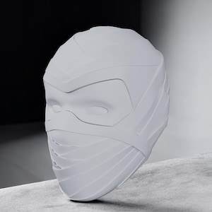 Pode incluir: Uma máscara cinza claro e estilizada com um design que cobre o rosto. A máscara apresenta aberturas para os olhos, uma ponte nasal e uma série de faixas horizontais que envolvem a cabeça. A forma geral é suave e arredondada, com uma estética futurista.