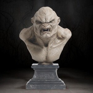May include: A detailed bust of a monstrous creature with a fierce expression. The sculpture features a snarling mouth with sharp teeth, furrowed brow, and a muscular chest. It is displayed on a grey pedestal, set against a dark background.