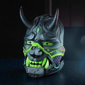May include: A black and green helmet with a demon-like design. The helmet features two black horns, green accents around the eyes and mouth, and a detailed face. The design has a futuristic, mechanical aesthetic.