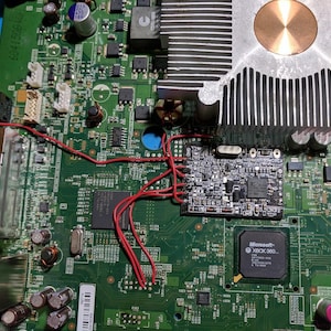 Könnte beinhalten: Nahaufnahme einer grünen Leiterplatte mit verschiedenen elektronischen Bauteilen. Rote Drähte verbinden mehrere Komponenten. Ein Prozessor mit der Aufschrift "Microsoft XBOX 360" ist zu sehen. Ein silberner Kühlkörper und eine kleine schwarze Platine sind ebenfalls vorhanden.
