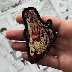Puede incluir: Parche bordado que representa un personaje de estilo anime con pelo largo rosa y un atuendo rojo. El parche tiene un borde negro, sobre un fondo de páginas de manga.