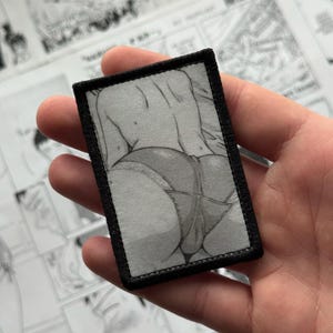 May include: A rectangular patch with a black border featuring a grayscale illustration of a person's lower back and buttocks. The illustration is detailed and suggestive, with visible shading and lines.