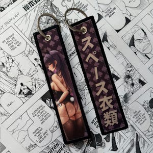 May include: Two rectangular keychains with a dark purple background and a repeating spade pattern. One keychain features a manga-style illustration of a woman in a bunny outfit. The other keychain has Japanese text.