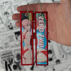 Puede incluir: Dos llaveros rectangulares rojos con borde rojo. Uno presenta un personaje de anime en traje de baño rojo, el otro el texto "ZERO TWO" y una vista parcial del personaje. Ambos tienen llaveros plateados.