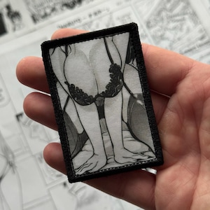 May include: A rectangular patch with a black border features a grayscale illustration of a person in lingerie. The design includes a bra and legs, with details in black and white. The patch is held in a hand.
