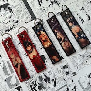Op de afbeelding: Vijf sleutelhangers met anime-stijl illustraties van vrouwen in verschillende outfits. De sleutelhangers hebben een rechthoekige vorm met een metalen ring aan de bovenkant. De achtergronden zijn zwart, rood en paars. De sleutelhangers worden weergegeven op een stripboekpagina.