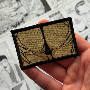Op de afbeelding: Geborduurde patch met een gouden achtergrond en een zwarte omtrek van een figuur. De patch heeft een zwarte rand. Het ontwerp is een gestileerde, abstracte weergave. De patch is rechthoekig.
