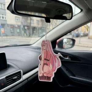 Puede incluir: Un ambientador para coche con una ilustración de estilo anime de una mujer con pelo largo rosa, vestida con lencería negra. El ambientador cuelga del espejo retrovisor de un coche.