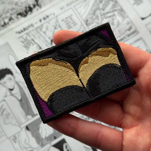 May include: A rectangular embroidered patch featuring a black background with two symmetrical gold-colored shapes that resemble stylized wings or curved forms. The edges of the patch are finished with a black border and small purple accents.
