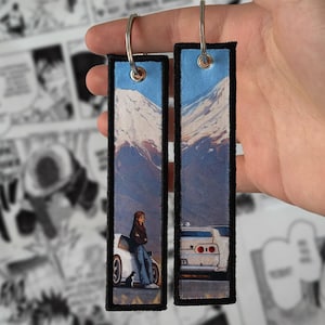 May include: Two rectangular fabric keychains with black stitched borders and metal rings. The design features a printed illustration of a person leaning against a white sports car with a snow-capped mountain in the background and a small cat nearby.
