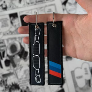 May include: Two black keychains with silver metal rings. One keychain features a white outline of a car part. The other has a design of blue, red, and black stripes. The keychains are rectangular and made of fabric.