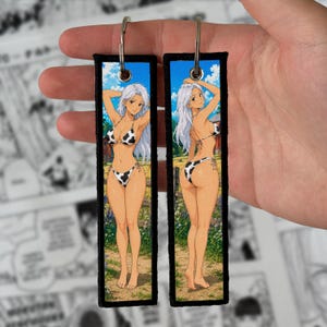 Puede incluir: Dos llaveros rectangulares de tela con un personaje de estilo anime de pelo largo blanco que lleva un bikini con estampado de vaca. El llavero izquierdo muestra una vista frontal y el derecho una vista trasera del personaje en un paisaje rural.