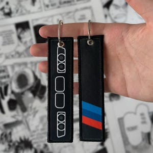Peut inclure: Deux porte-clés rectangulaires noirs avec des anneaux argentés. L'un des porte-clés présente un contour blanc de l'avant d'une voiture, tandis que l'autre a des rayures bleues, rouges et noires. Les porte-clés sont tenus.