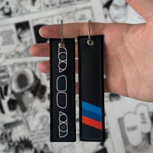 May include: Two rectangular black fabric keychains with metal rings. One keychain features white embroidered outlines of car headlights and a grille. The other keychain displays a diagonal stripe pattern in blue, light blue, and red colours.