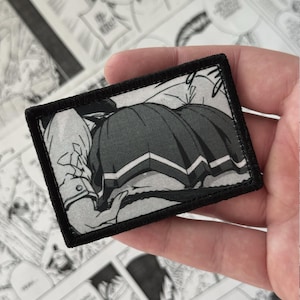 Op de afbeelding: Een rechthoekige geborduurde patch met een zwart-witte manga-illustratie van een personage dat een plooirok draagt. De patch heeft een dikke zwarte gestikte rand en een gedetailleerde grafische print van kledingstukken.