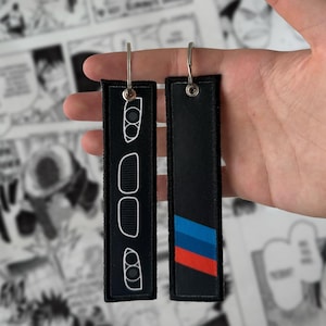 Puede incluir: Dos llaveros rectangulares negros con aros de metal plateado. Un llavero presenta un contorno blanco de piezas de automóvil. El otro tiene rayas diagonales azules, rojas y negras. El fondo es una página de cómic.