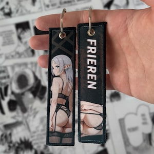 Op de afbeelding: Twee zwarte rechthoekige sleutelhangers met zilveren metalen ringen. Eén sleutelhanger heeft een cartoonillustratie van een vrouw in lingerie, de andere heeft het woord "FRIEREN" in het wit. De sleutelhangers worden in de hand van een persoon gehouden.