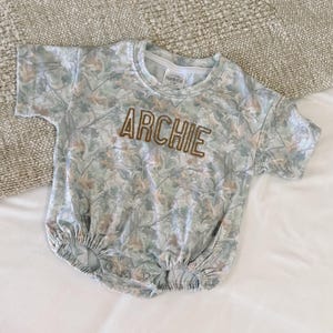 May include: A light blue baby romper with a leaf pattern and the name "ARCHIE" embroidered in brown thread. The short-sleeved romper has a round neckline and snap closures at the bottom.
