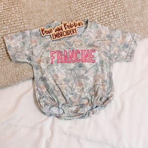 May include: A short-sleeved baby romper featuring a camouflage pattern of muted green, brown, and grey leaves. The name FRANCINE is embroidered in bright pink thread across the chest. A wooden sign reading Bow and Bobbin EMBROIDERY is placed above.