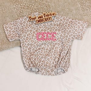 May include: A short-sleeve romper featuring a tan and brown leopard print pattern. The center of the chest displays the name CECE embroidered in bright pink thread. A wooden sign with the text Bow and Bobbin EMBROIDERY rests on the neckline.