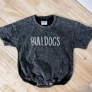 May include: A charcoal gray acid-wash short-sleeve romper featuring the word BULLDOGS printed in white capital letters across the chest. The garment has a round neckline, short sleeves, and a snap-button closure at the bottom crotch area.