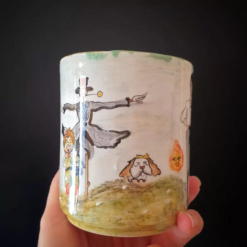 May include: A white ceramic mug featuring a hand-painted illustration of characters from a fantasy film. The design includes a scarecrow-like figure, a dog with a crown, and a small flame. The mug is held by a hand.
