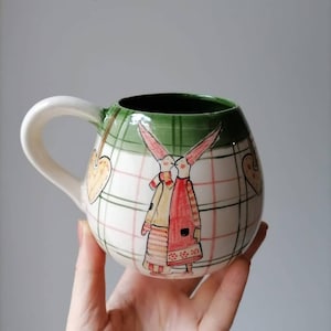 Puede incluir: Una taza de cerámica con asa blanca y cuerpo redondeado. La taza presenta un borde verde y un diseño de dos conejos con abrigos y bufandas, con un patrón de corazón y cuadros. La taza está sujeta por una mano.