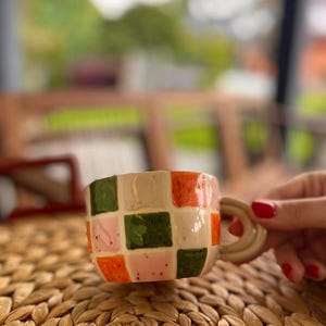 Może przedstawiać: Ceramiczny kubek z kremową podstawą i kolorowym wzorem szachownicy z pomarańczowych, zielonych i różowych kwadratów. Kubek ma zaokrąglony uchwyt i jest ustawiony na tkanej powierzchni.