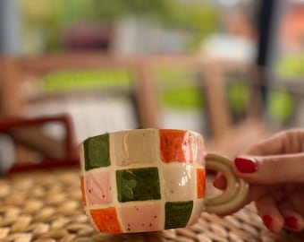 May include: A ceramic mug with a cream-colored base and a colorful checkerboard pattern of orange, green, and pink squares. The mug has a rounded handle and is set on a woven surface.