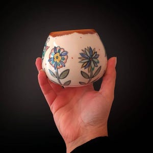 May include: A small, round, off-white ceramic bowl with a brown rim. The bowl is decorated with hand-painted flowers in blue, yellow, and orange. The bowl is held in a hand against a black background. The bowl is approximately 3 inches in diameter.