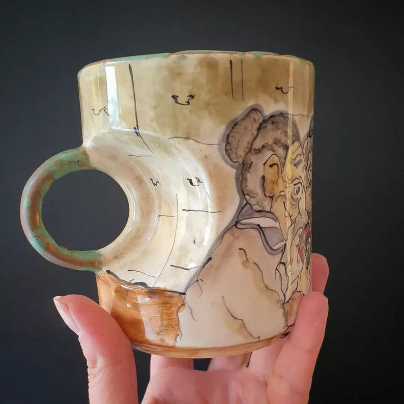 May include: Hand-painted ceramic mug with a unique handle design. The mug features a beige background with a painted illustration of figures. The handle is green and the base is brown. The mug is held by a hand.