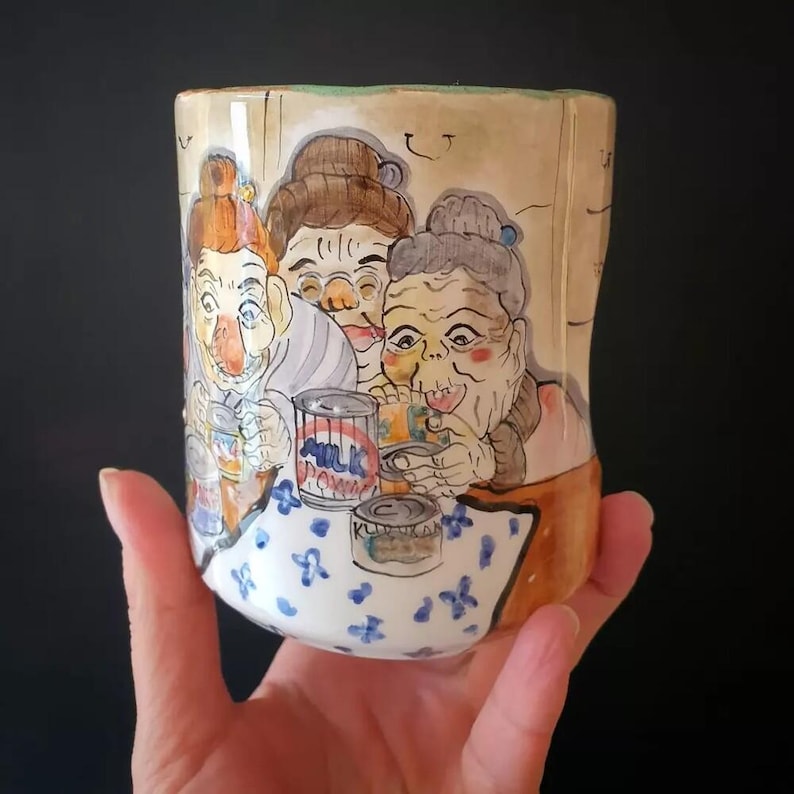 May include: A hand-painted ceramic mug featuring a whimsical illustration of three elderly women. The artwork includes details like a table with blue floral patterns and cans of milk. The mug is held by a hand.