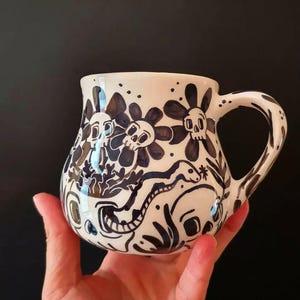 Peut inclure: Mug en céramique blanche avec un motif floral et crâne noir. La tasse a une anse incurvée et une base large. L'œuvre d'art comprend des fleurs stylisées, des crânes et un serpent. La tasse est tenue sur un fond noir.