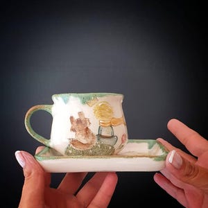 May include: A white ceramic teacup and saucer set with a hand-painted illustration of a fox and a young boy with blonde hair. The cup and saucer have a green trim and handle. The set is held by a person.