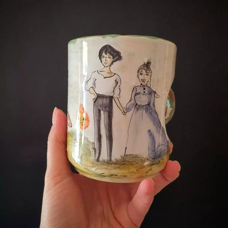 May include: A ceramic mug with a hand-painted illustration of two figures holding hands. The figures are dressed in white and blue clothing. The mug has a green rim and a yellow base.